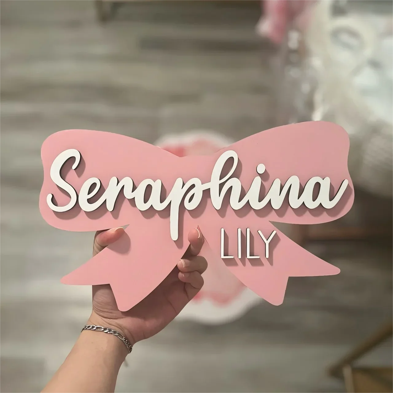 Pink wood baby name sign shaped like a bow with "Seraphina" and "Lily" in white text.
