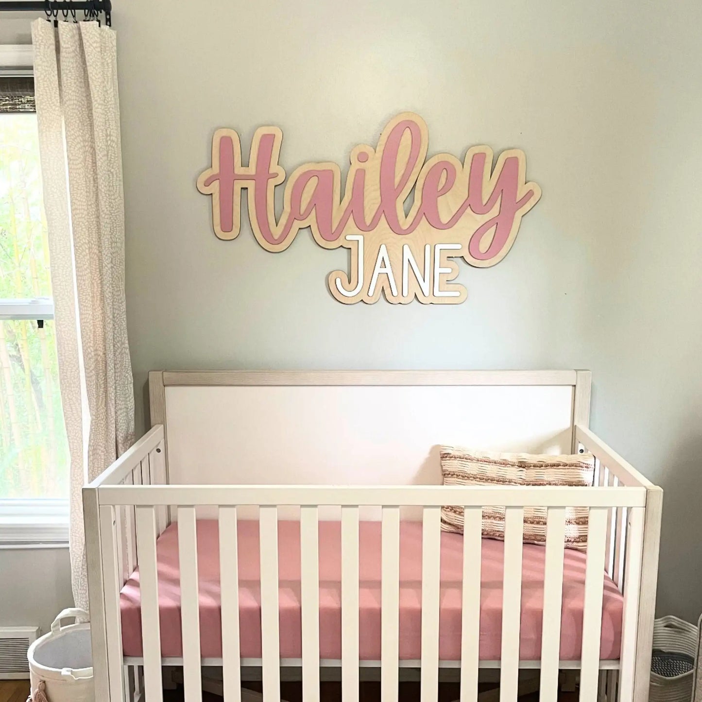 Customized wood baby name sign in pink and white above a crib in a nursery room.