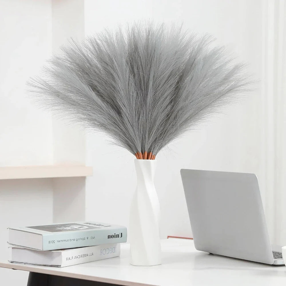 Artificial pampas grass in a white vase on a desk with books and laptop, featuring a fluffy, gray appearance.