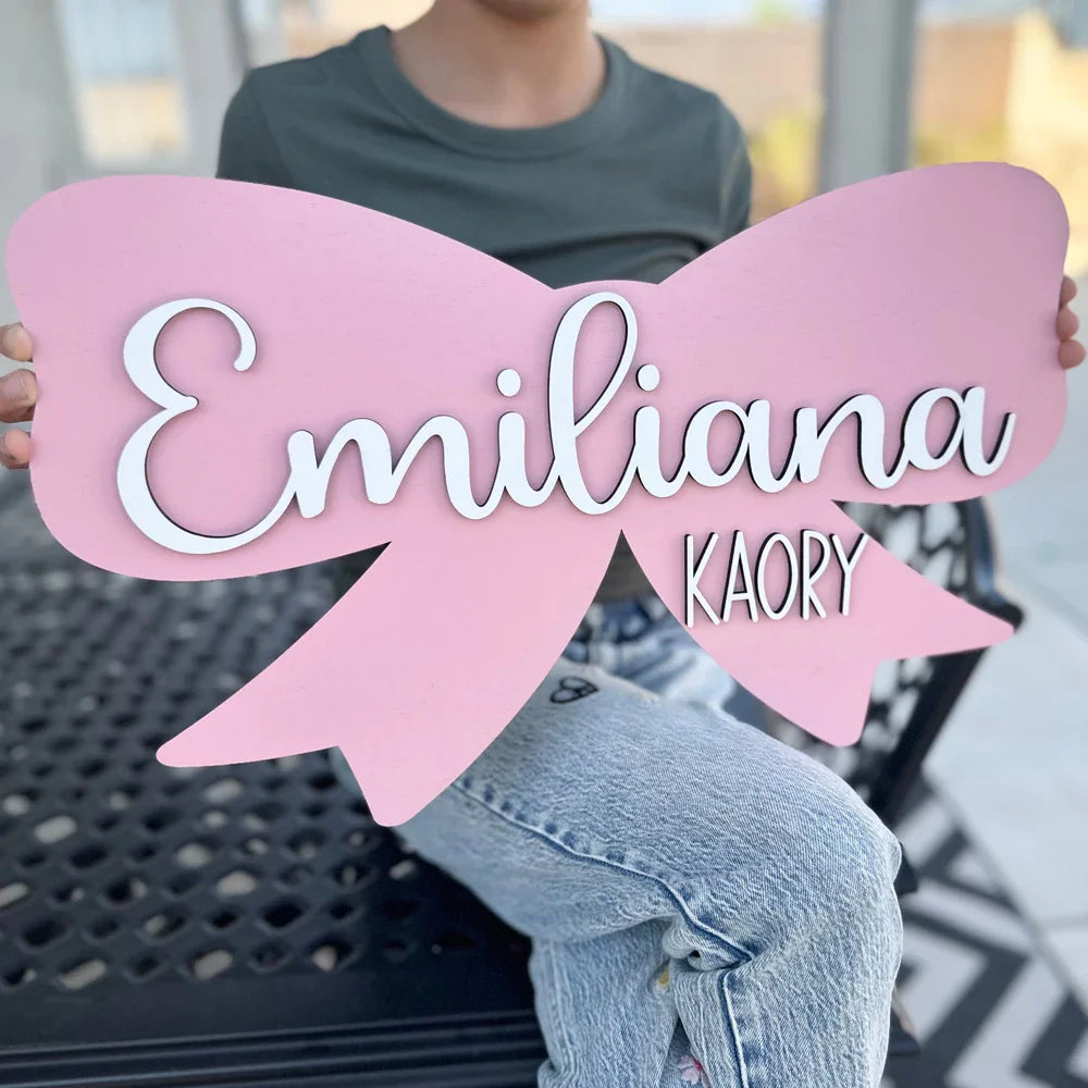 Pink bow-shaped wooden baby name sign displaying "Emiliana Kaory" for nursery wall decor.