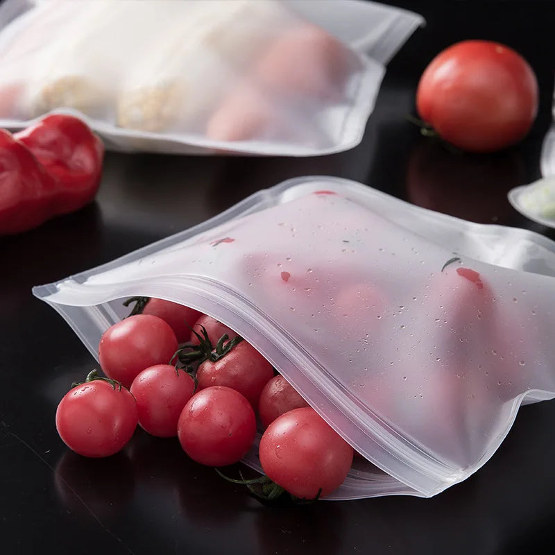 Reusable silicone food storage bags with zip seal containing cherry tomatoes and vegetables on a black surface.