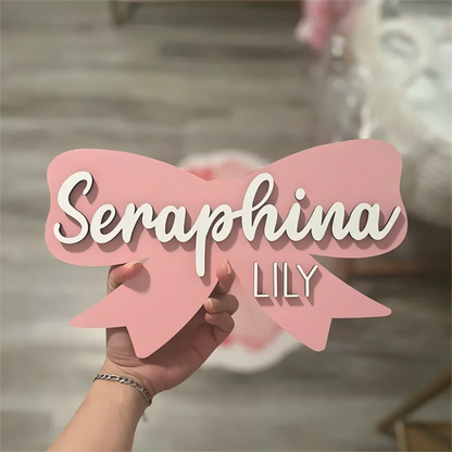 Pink wood baby name sign shaped like a bow with "Seraphina" and "Lily" in white text.