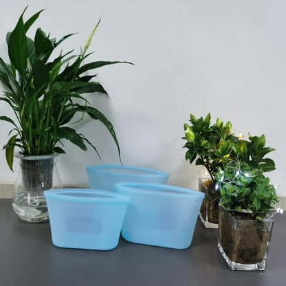 Three blue reusable silicone food storage bags on a kitchen counter with plants.