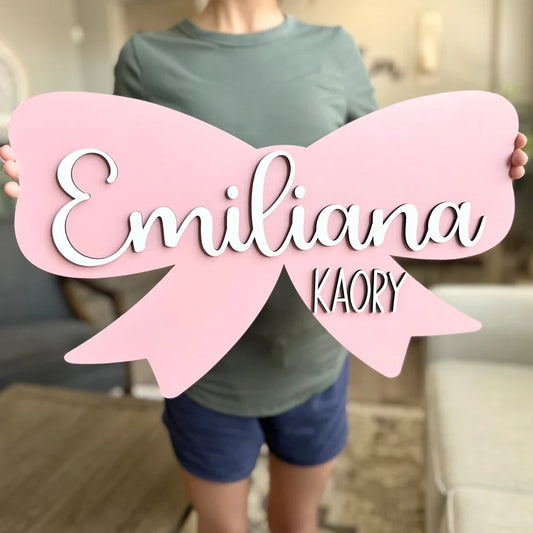 Custom wooden baby name sign in pink bow shape with "Emiliana Kaory" text for nursery wall decor.