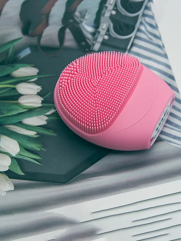 Pink electric facial cleansing brush with silicone bristles on a decorative surface.